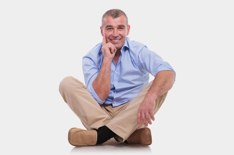 casual senior man sitting on the floor with his legs crossed and holding his hand at his chin, in a pensive way, while smiling for the camera. isolated on white background; Shutterstock ID 141171721; Department: -
