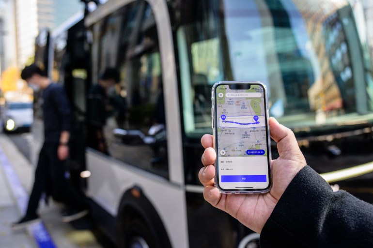 This photo taken in Seoul on November 23, 2022 shows a person posing with a phone to show the app for the country's first self-driving bus route run by 42 Dot, a start-up owned by South Korea's Hyundai which created the automomous driving technology. - South Korea's capital launched its first self-driving bus route on November 25, part of an experiment which engineers said aims to make the public feel more comfortable with driverless vehicles on the roads. (Photo by Anthony WALLACE / AFP)