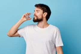 A man in a t-shirtشرب 8 أكواب من الماء يوميا.. حقيقة أم خرافة؟ and trousers on a blue background drinking water from a glass ; Shutterstock ID 1261170649; purchase_order: multimedia; job: ; client: ; other: