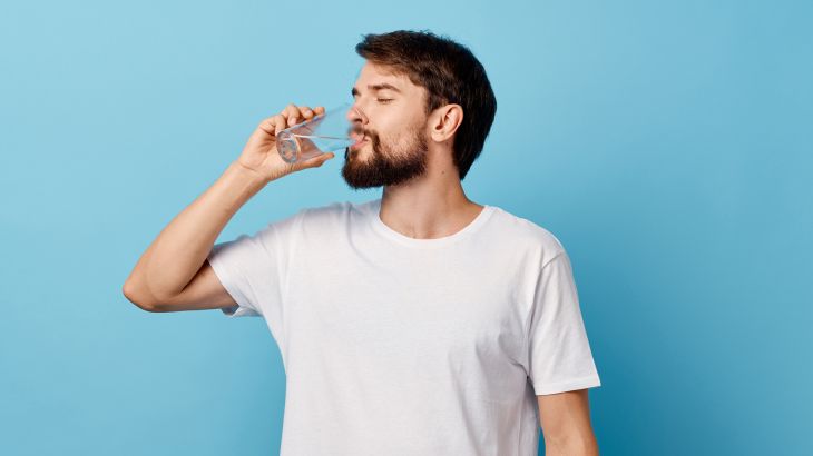 A man in a t-shirtشرب 8 أكواب من الماء يوميا.. حقيقة أم خرافة؟ and trousers on a blue background drinking water from a glass ; Shutterstock ID 1261170649; purchase_order: multimedia; job: ; client: ; other:
