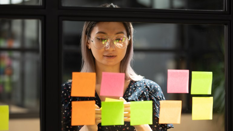 Komoly összpontosított fiatal női edző tanár diák ázsiai üzletasszony dolgozik projekt stratégia terv írás cél feladatok kreatív ötletek ragadós post jegyzetek üveg scrum board irodai fal