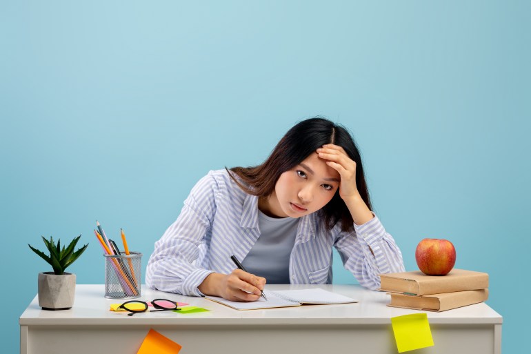 Tiresome task. Exhausted asian lady sitting at table, writing in notebook, leaning head on hand, studying over blue background. Sad and tired female student feeling headache, looking at camera