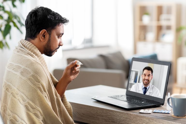 healthcare, technology and people concept - sick indian man in blanket with thermometer having video call or online consultation with smiling doctor on laptop computer at home
