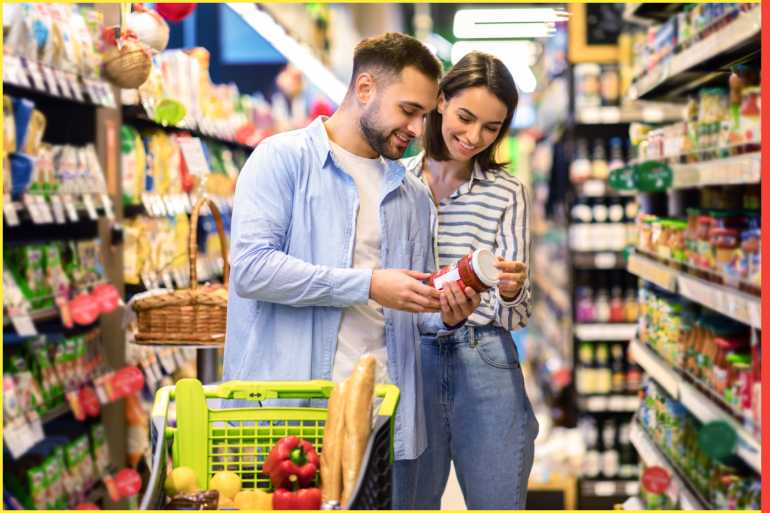 Happy Couple Buying Food In Supermarket, Choosing Products Standing With Trolley Cart Along Aisles And Full Shelves Purchasing Groceries Essentials Together. Smiling Spouses Holding Jar Of Sause