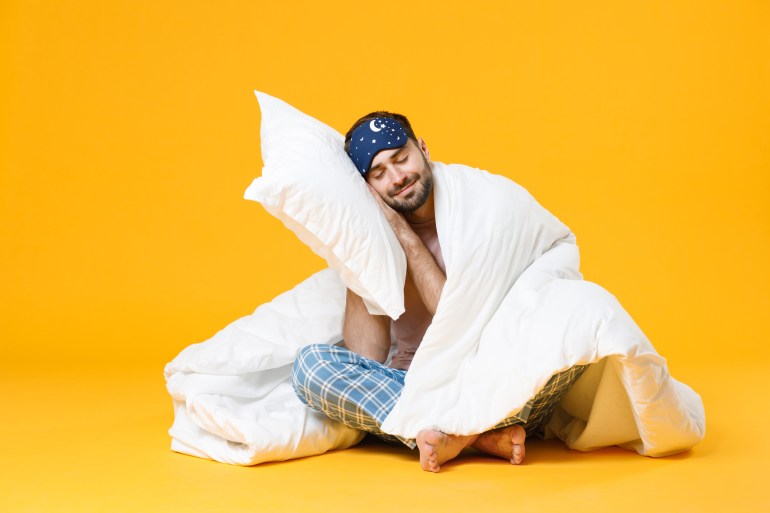 Full length of smiling young bearded man in pajamas home wear sleep mask sitting with pillow blanket isolated on bright yellow colour background studio portrait. Relax good mood lifestyle concept; Shutterstock ID 1885457752; purchase_order:aja; job:; client:; other: