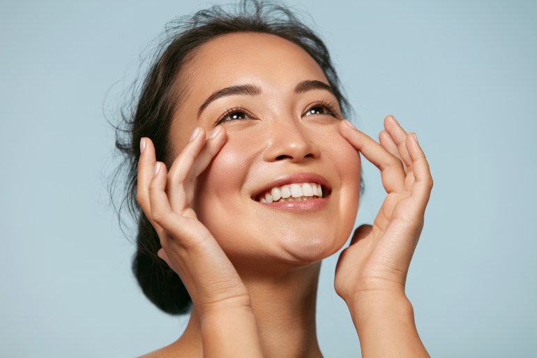 Skin care. Woman with beauty face touching healthy facial skin portrait. Beautiful smiling asian girl model with natural makeup touching glowing hydrated skin on blue background closeup; Shutterstock ID 1543602947; purchase_order: ajnet; job: ; client: ; other: