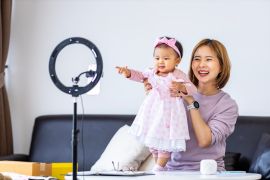 Young Asian influencer mother is selling children clothes live online streaming at home while raising her toddler as assistance