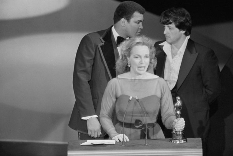 Actress Beatrice Straight makes her acceptance speech for her Best Supporting Actress Oscar for her role in Network, as actor Sylvester Stallone and world heavyweight boxing champion Muhammad Ali stare each other down behind her. Ali made a surprise appearance on stage as Stallone was making the announcement that Straight had won the award at the 49th Annual Academy Award presentation at the Music Center, and for a few moments playfully sparred on stage.