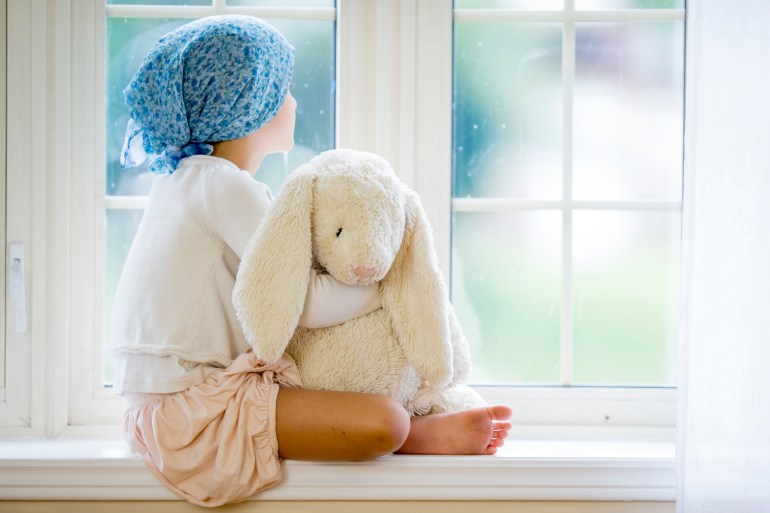 A little girl with cancer is sitting by the window and is looking outside. She is holding a stuffed animal.