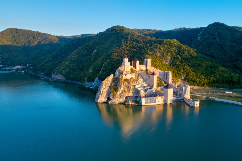 The medieval fortress of Golubac, mirroring in the waters of the Danube. Fortress towers illuminated by warm sunset light, blue sky. Aerial shot. Famous tourist place, Serbia.