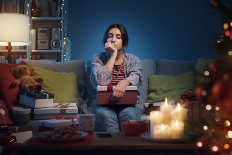 Disappointed woman opening Christmas gifts at home, she is lonely and sad; Shutterstock ID 2221787415; purchase_order: aljazeera ; job: ; client: ; other: