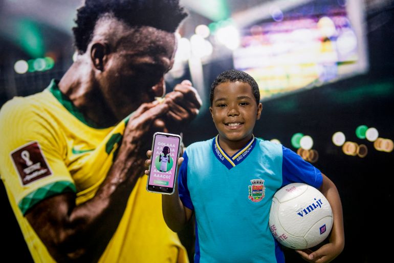 Pupil Yuri Rodrigues da Silva poses for a picture with a mobile phone showing the Vini Jr Institute educational app at the Visconde de Sepetiba municipal school in Sao Goncalo, on the outskirts of Rio de Janeiro, Brazil, on February 27, 2024. - The Vini Jr Institute, founded by Real Madrid's Brazilian forward Vinicius Junior, aims to improve public education in disadvantaged communities in Brazil through football and technology. Some 4,500 students and 500 teachers in four Brazilian states have so far benefited from the project launched in 2021. (Photo by Daniel Ramalho / AFP)