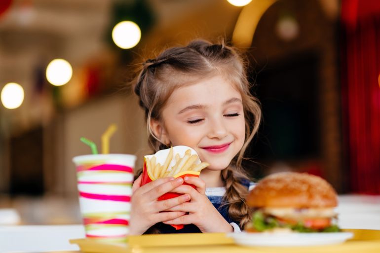 Little girl eating hamburger and French fries in a fast food restaurant. Child having sandwich and potato chips for lunch. Kids eat unhealthy fat food. Grilled fastfood sandwich for children.